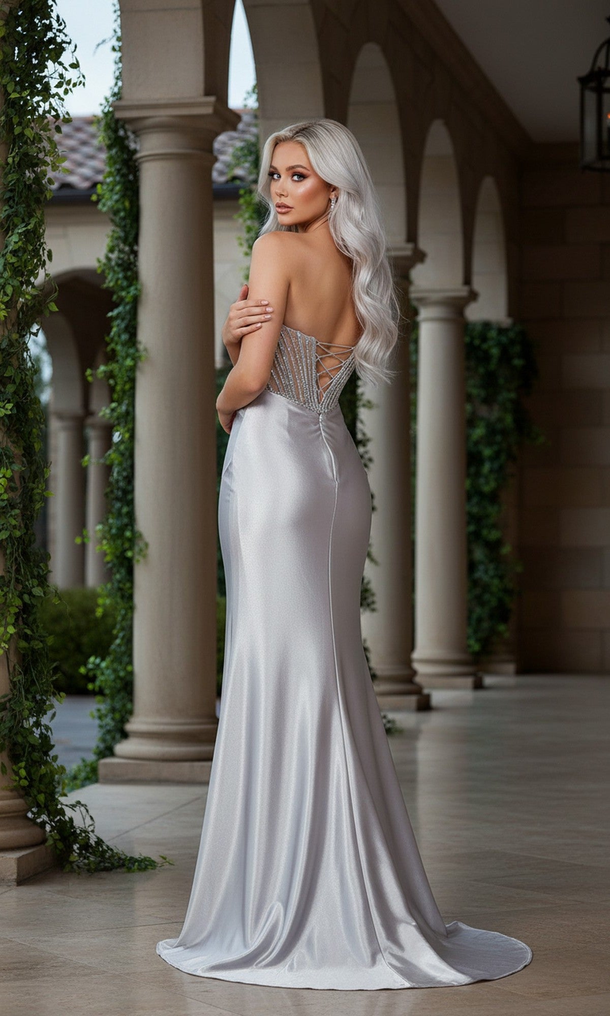 A woman with long, silver hair stands in an arched corridor wearing the Cinderella Couture 8473J, a silver satin long prom dress with a sheer corset bodice and lace-up detailing.